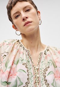 Λουλουδάτη μπλούζα σε ελαφρύ ύφασμα με κρεμ χρώμα, διακόσμηση με ροζ λουλούδια, πράσινα φύλλα και πολύπλοκες λεπτομέρειες, κλείσιμο με κουμπιά και φουσκωτά μανίκια.