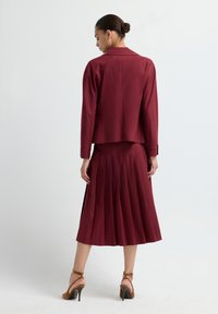 Burgundy skirt suit with pleated skirt, fitted jacket, notched collar, long sleeves, and pointed heels. Smooth texture, sophisticated design.
