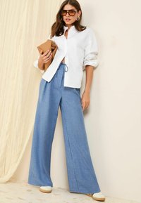 Camisa blanca de botones, pantalones de lino azul anchos y zapatos crema. Los accesorios incluyen gafas de sol oversized y un bolso de mano marrón.