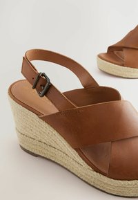 Brown leather wedge sandals with crisscross straps, an adjustable ankle strap, and a woven jute midsole. Smooth texture and rounded toe.