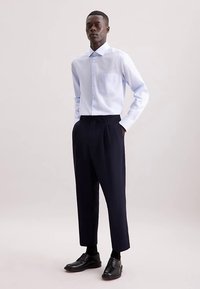 Camicia a righe azzurro chiaro con colletto appuntito, taschino sul petto singolo, abbinata a pantaloni navy plissettati e scarpe nere.