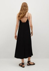 Černé midi šaty s tenkými ramínky a designem ve tvaru V na zádech. Vyrobeny z měkké tkaniny s plynulou siluetou. Doplňují je ploché sandály s pásky.