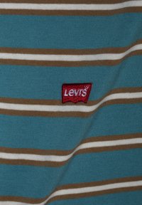 Modrá pruhovaná tkanina se hnědými a bílými pruhy. Obsahuje červeně vyšívané logo "Levi's" v dolním levém rohu.