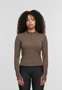 Braunes langärmliges Oberteil mit einem Viertel-Reißverschluss, taillierter Passform und strukturierten Nähten. Hergestellt aus glattem Stoff; kombiniert mit schwarzen hochgeschnittenen Leggings.