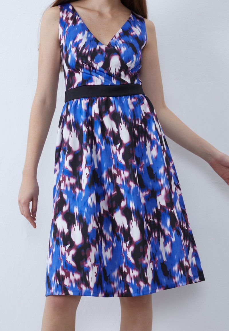 Robe sans manches longueur genou avec col en V, motif abstrait bleu, noir, blanc et violet, et une ceinture noire cintrée sur une femme debout.