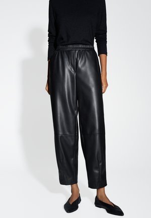 Frau trägt hoch taillierte, schwarze Lederhose mit weitem Bein, schwarzen Langarmoberteil und schwarze spitze Ballerinas vor weißem Hintergrund.