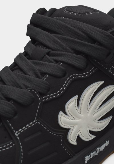 Κοντινό πλάνο μαύρου αθλητικού παπουτσιού με χοντρές μαύρες κορδόνια, λευκό ραμμένο λογότυπο που μοιάζει με στυλιζαρισμένο φύλλο και το κείμενο «palm angels» στη σόλα.