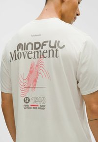 Homme portant une chemise beige clair avec le motif « Mindful Movement », une vague rouge, « 1998 » et le texte « Trouvez le flux dans le voyage » dans le dos.