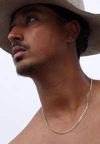 Eine mit Gold überzogene Kette liegt um den Hals; kurze, flache Glieder; der Träger trägt einen hellgrauen Hut mit breiter Krempe. Die Hautfarbe ist warm und hat Gesichtshaar.