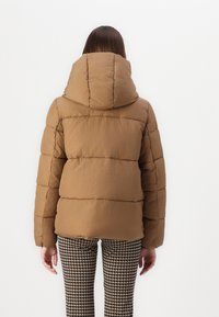 Giacca puffer marrone con cappuccio, caratterizzata da una texture trapuntata e cuciture orizzontali. Abbinata a pantaloni a scacchi bianchi e neri.