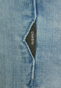 Denim tkanina u svijetloplavoj boji s blagom teksturom. Na bočnoj šavu je pričvršćena crna oznaka s natpisom "CHASIN'".