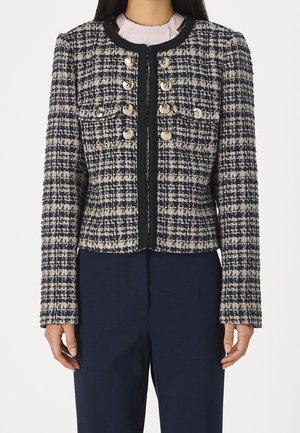 Nainen, joka käyttää laivastonsinistä ja beigen väristä tweed-takkia, jossa on koristeelliset hopeiset napit ja musta reunus, laivastonsinisten housujen päällä.