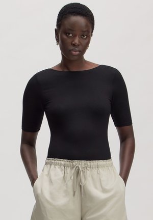 Vrouw met kort haar, gekleed in een zwarte aansluitende top en beige broek met trekkoord, staand met handen in de zakken tegen een rustige achtergrond.