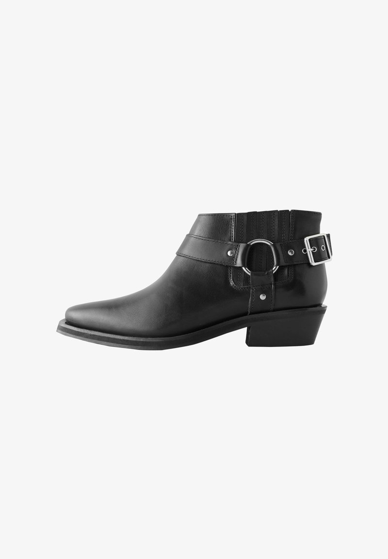 Black leather ankle boot with a pointed toe, featuring a wide strap, circular hardware, a buckle, and textured elastic panels on the back.
