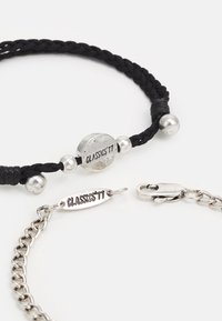 Pulseira trançada preta com bead redondo prateado rotulado "CLASSICS'77," emparelhada com uma pulseira de corrente prateada com fecho e etiqueta.