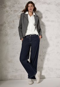 Grijze cropped jasje, witte overhemden, donkerblauwe wijde jeans en witte sneakers. Gladde texturen met contrasterende kleuren en naden.