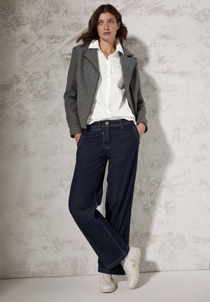 Giacca grigia corta, camicia bianca con bottoni, jeans larghi blu scuro e sneakers bianche. Texture lisce con colori e cuciture a contrasto.