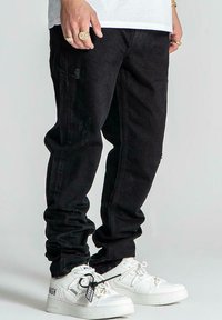 Jeans de mezclilla negra con un corte ajustado, que presentan una textura desgastada. Combinados con zapatillas deportivas blancas y detalles decorativos metálicos visibles.
