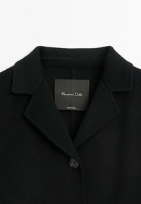 Manteau en laine noire avec col à revers cranté et fermeture par un seul bouton noir, avec l'étiquette de la marque Massimo Dutti à l'intérieur du col.