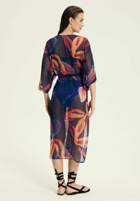 Przezroczysty, granatowy narzut na ubranie z żywymi, wielokolorowymi abstrakcyjnymi wzorami. Ma rękawy do trzech czwartej oraz wiązanie w talii. Czarne sandały na paskach.