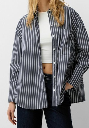 Woman wearing open navy blue and white vertical striped button-up shirt over a white crop top and dark jeans, hand in pocket.