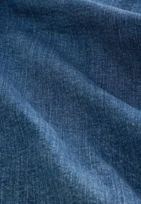 Close-up van gestructureerde blauwe spijkerstof met zichtbaar weefpatroon en lichte kreukels die zachte plooien vormen.