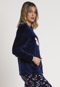 Sudadera de felpa azul marino con un diseño de animal rosa, que incluye un bolsillo frontal y una textura suave, combinada con pantalones de pijama azul marino con un estampado.
