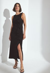 Musta mekko, jossa on röyhelömuotoilu sivussa ja reiden yläpuolelle ulottuva halkio. Kangas on sileä ja siluetti istuva. Yhdistetty litteisiin sandaaleihin.