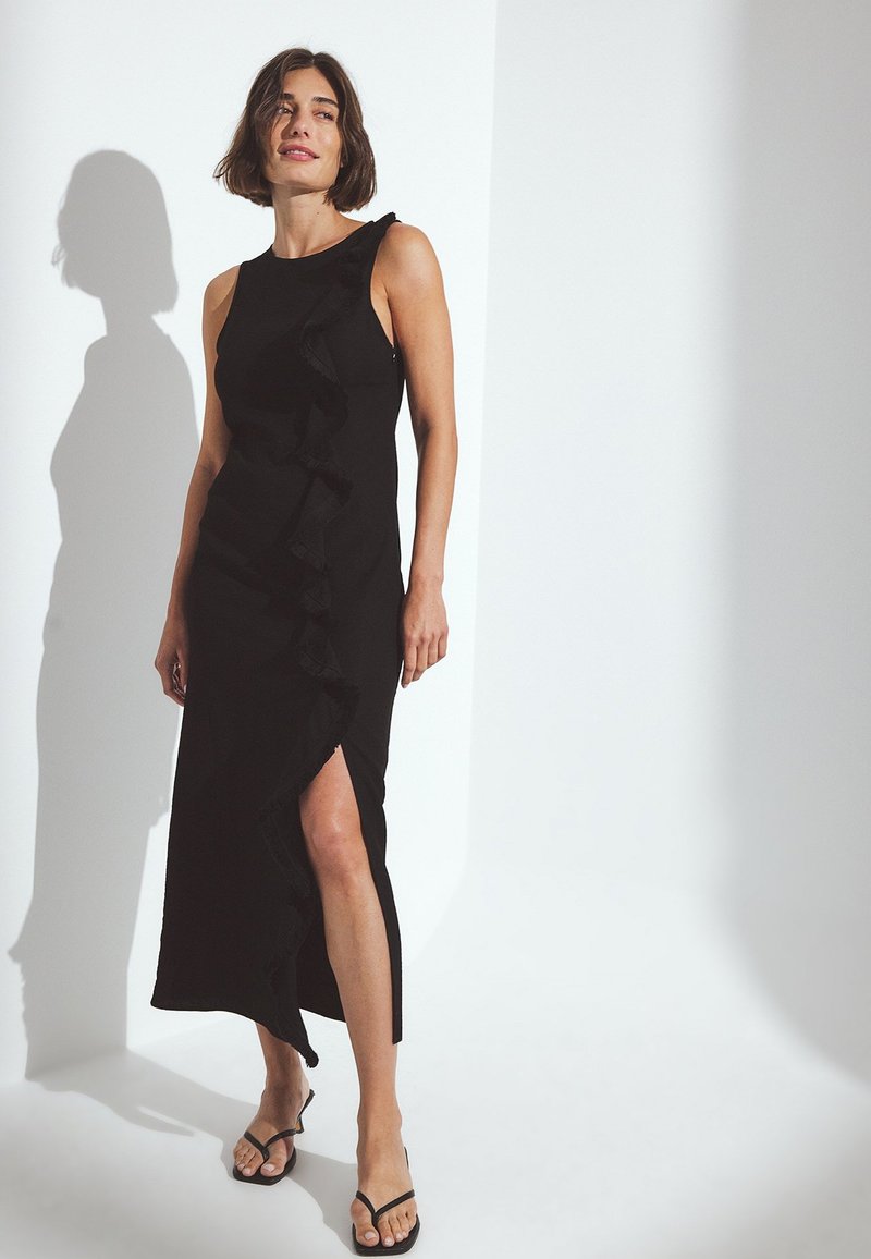 Musta mekko, jossa on röyhelömuotoilu sivussa ja reiden yläpuolelle ulottuva halkio. Kangas on sileä ja siluetti istuva. Yhdistetty litteisiin sandaaleihin.