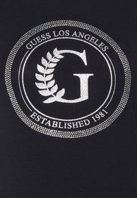 Sort T-shirt med et sølvfarvet cirkulært grafisk design med teksten "Guess Los Angeles" og et laurbærdesign, prydet med rhinsten.