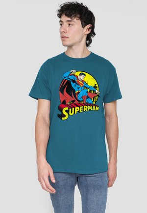 Young man wearing teal t-shirt with vintage Superman graphic in flying pose over a yellow circle, paired with blue jeans.