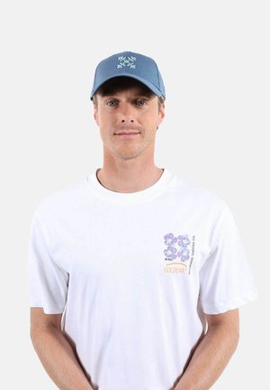 Homme portant une casquette bleue à motif floral et un t-shirt blanc avec des fleurs violettes et le texte "OXBOW Authentic Surfing Co.".