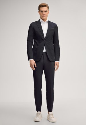 Jeune homme debout portant un costume noir ajusté avec une chemise blanche et des baskets blanches sur un fond clair uni.