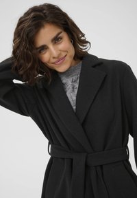 Manteau en laine noire mélangée avec une taille ceinturée, revers crantés et manches longues, porté sur un pull en tricot gris avec un motif texturé.