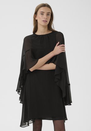 Hosszú barna hajú nő fekete, áttetsző réteges ruhában, hosszú ujjakkal, karját keresztbe téve áll fehér háttér előtt.