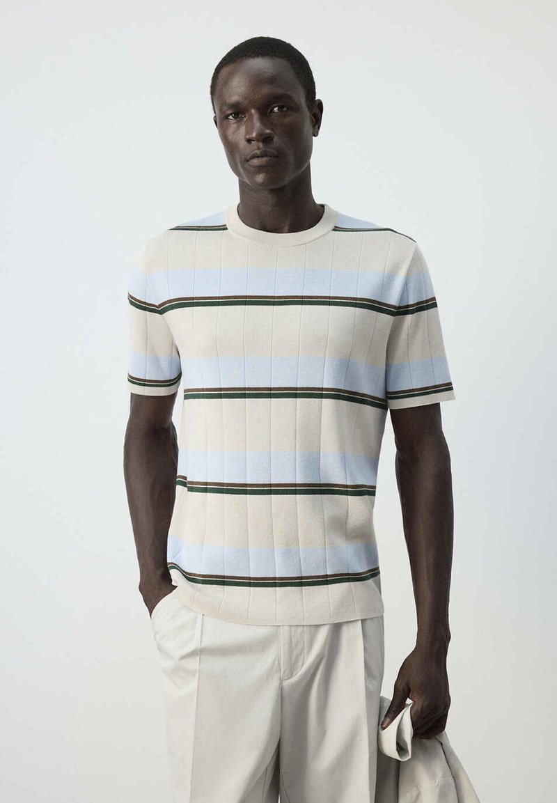 Homme portant un pull à manches courtes rayé beige, bleu et vert et un pantalon clair, tenant une veste de couleur claire sur un fond uni.