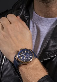 Modrý chronograf s textúrovaným tmavomodrým koženým remienkom a zlatými akcentmi, ktorý má okrúhly ciferník so striebornými označeniami a subciferníkmi.