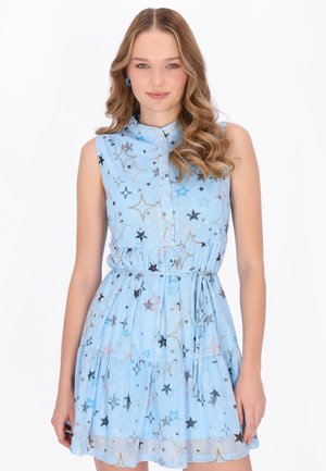 Młoda kobieta o falowanych włosach, ubrana w bezrękawnikową, jasnoniebieską sukienkę w gwiazdy, z taliowaniem, stojąca na białym tle.