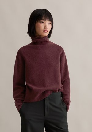 Burgundy turtleneck sweater made of soft, textured fabric with loose fit. Paired with black trousers, featuring side pockets.