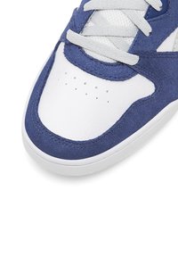 Tuvumā redzama baltā sporta apava purngala daļa ar zilu zamšādu, perforācijas caurumiem un baltām šņorēm uz baltas fona.