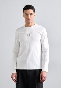Hvid langærmet t-shirt lavet af bomuld, med et lille A/X-logo i lys brun på brystet og rund halsudskæring, parret med mørke bukser.