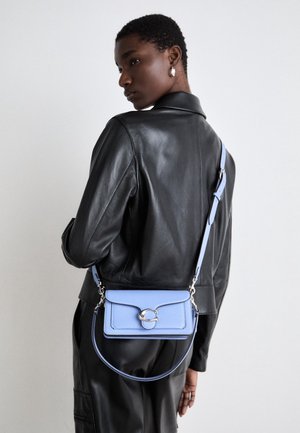 Personne portant une veste et un pantalon en cuir noir, portant un sac bandoulière bleu clair avec une boucle argentée, vue de profil sur un fond uni.
