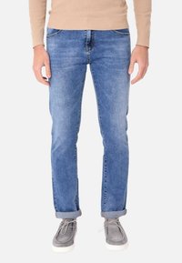 Jeans in denim blu da uomo con vestibilità slim, caratterizzati da un lavaggio chiaro, un leggero scolorimento e risvolti, abbinati a scarpe casual grigie.