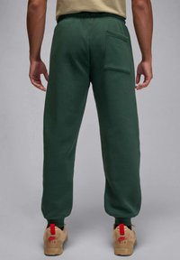 Grüne Sweatpants mit elastischem Bund, einer einzelnen Gesäßtasche, legerer Passform und weicher Textur, kombiniert mit beigen Sneakers mit roten Akzenten.