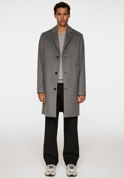 Uomo che indossa un cappotto grigio con bottoni sopra un maglione beige, pantaloni neri e sneakers bianche, in piedi contro uno sfondo chiaro semplice.