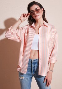 Prúhovaná košeľa s gombíkmi v broskyňovej a bielej farbe s dlhými rukávmi, náprsnými vreckami a voľným strihom, nosená cez biely crop top a modré džínsy.