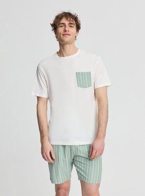 Giovane uomo che indossa una camicia bianca a maniche corte con tasca a righe verdi e pantaloncini abbinati a righe verdi, in piedi davanti a uno sfondo semplice.