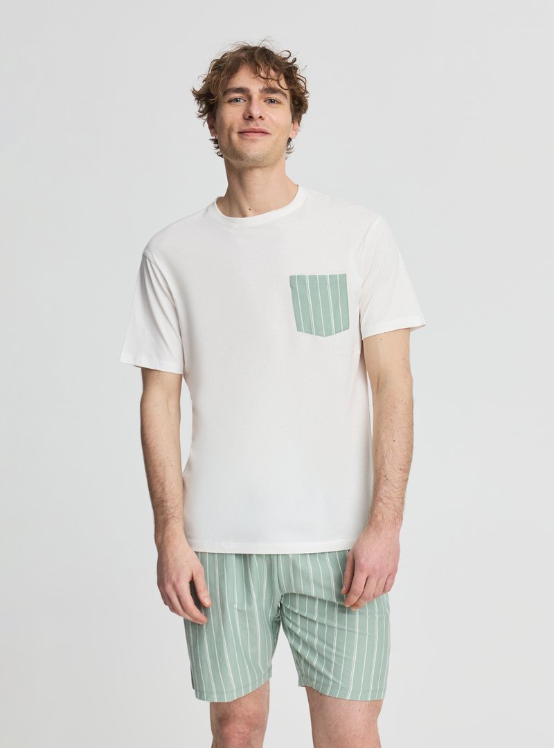 Młody mężczyzna ubrany w białą koszulę z krótkim rękawem z zieloną paskowaną kieszenią i pasujące zielone paskowane szorty, stojący na jednolitym tle.