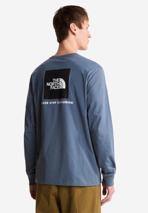 Mężczyzna w niebieskiej koszuli z długim rękawem z logo "The North Face" oraz napisem "Never Stop Exploring" na plecach, w połączeniu z brązowymi spodniami.