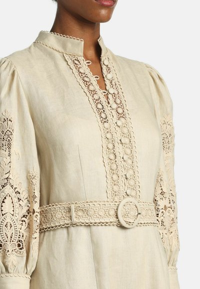 Beige jurk met pofmouwen met ingewikkelde kanten inzetten, stof bedekte knopen aan de voorkant en een bijpassende riem met ronde gesp.